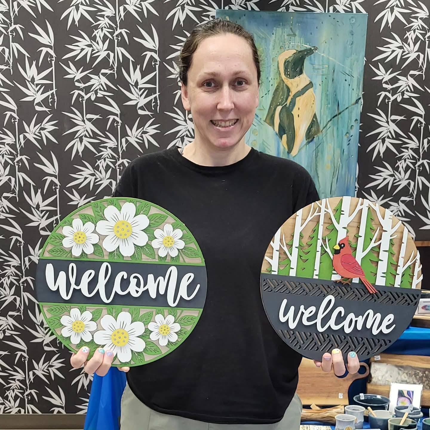 Katie Holmes, RMT, holding up custom signs made by Brandon Johns for the spring 2026 paint night at Wrobel Massage Therapy.